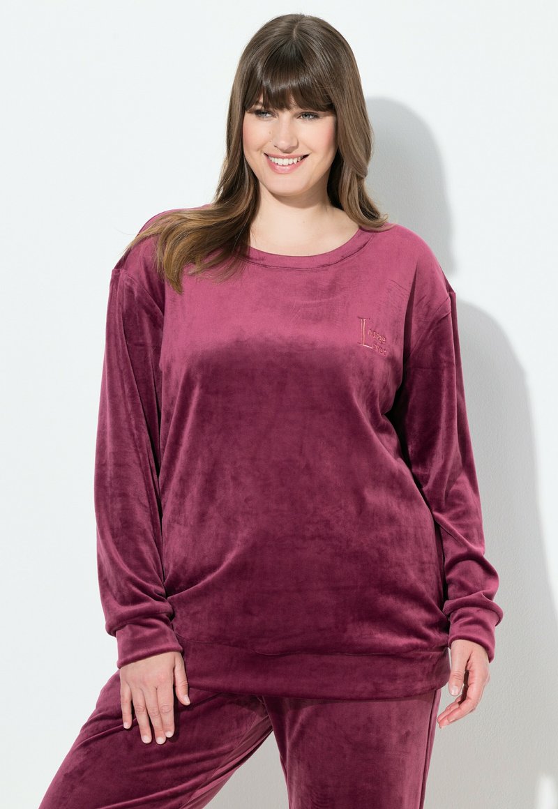 Donna sorridente con capelli castani che indossa un top viola in velluto a maniche lunghe e larghe, abbinato a pantaloni, su uno sfondo chiaro e neutro.