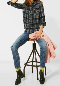 Marine-rutete knap skjorte med grønne og cremefarvede accenter, denimjeans med slidte detaljer, sorte ankelstøvler og en lys pink jakke.
