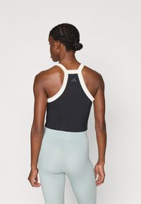 Moteris su nertomis kasomis, vilkinti juodą bepečių viršų su baltu apvadu ir šviesiai mėlynomis aukštai supjaustytomis leginsais, atsisukusi atgal neutralioje fone.