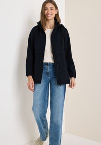 Veste marine avec fermeture éclair à l'avant, capuche réglable et poignets élastiques. Associée à un jean bleu clair à larges jambes et des baskets neutres.