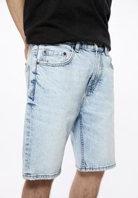 Vaaleansiniset farkkushortsit, joissa on suora leikkaus, viisi taskua ja metallinitteet. Ominaisuuksiin kuuluu revityt helmareunat ja haalistunut pesu kuluneen ilmeen saamiseksi.