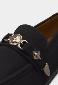 Μαύρο δερμάτινο μοκασίνι με ασημένιες διακοσμήσεις πεταλούδας και γεωμετρικά μεταλλικά στοιχεία στο λουράκι μπροστά.