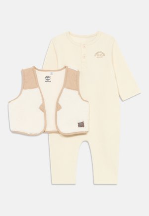 Combinaison à manches longues côtelée couleur crème avec patte de boutonnage, associée à un gilet doublé en polaire beige et blanc avec un écusson logo.
