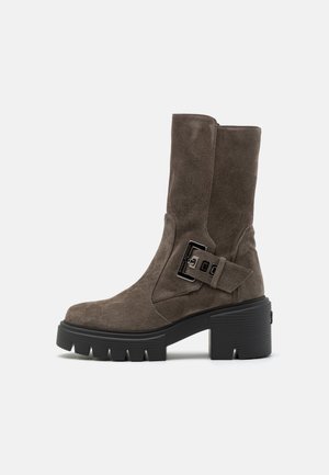 Brauner Wildleder-Stiefel mit mittlerer Schaftlänge, grober schwarzer Profilsohle, klobigem Blockabsatz und dekorativem seitlichem Schnallenriemen.