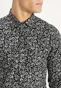 Uomo che indossa una camicia nera con un motivo floreale bianco e bottoni a pressione, dotata di due tasche sul petto e di una piccola etichetta "Replay" sulla tasca destra.