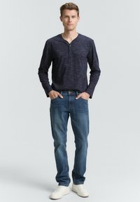 Marineblå langærmet henley-skjorte med et tekstureret design, parret med lys blå jeans og hvide sneakers, stående foran en neutral baggrund.