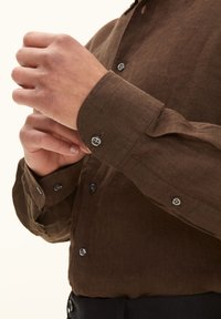 Camicia di lino marrone con maniche lunghe e polsini abbottonati. Presenta bottoni metallici e una vestibilità casual, mettendo in risalto un tessuto texturizzato.