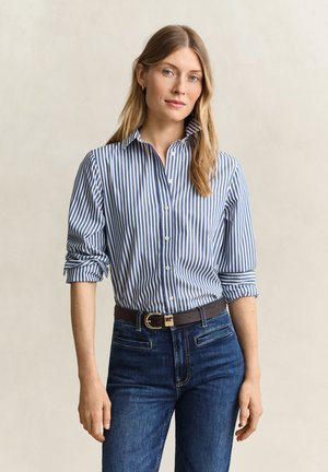 Femme aux longs cheveux blonds portant une chemise rayée bleue et blanche boutonnée avec les manches retroussées et un jean bleu foncé avec une ceinture marron.