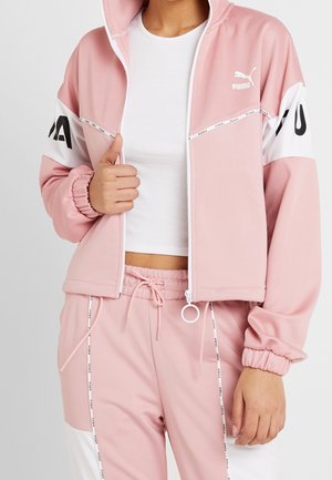 Persoon draagt een roze Puma trainingspak met witte en zwarte accenten, zichtbaar wit cropped topje, hand houdt de ritssluiting van het jasje vast.