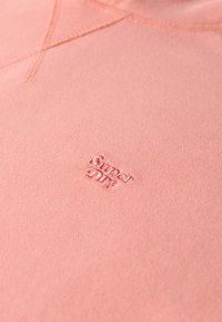 Sudadera rosa claro con tejido texturizado y detalles cosidos. Presenta un pequeño logo bordado de "Superdry" en hilo a juego.