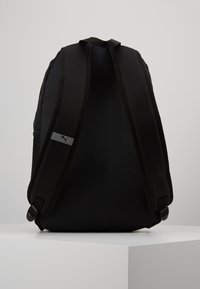 Zaino nero con schiena imbottita, spallacci doppi, caratterizzato da un piccolo tag con logo su una delle cinghie e una texture liscia opaca.