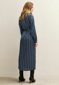 Vrouw met blond haar draagt een lange marineblauwe jurk met witte verticale strepen en zwarte hakken, staande tegen een effen muur.
