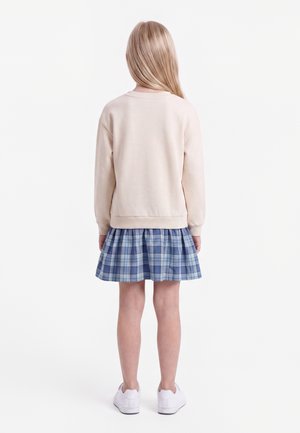 Enfant aux longs cheveux blonds portant un sweat-shirt beige, une jupe à carreaux bleue et des baskets blanches, debout de dos sur fond blanc.