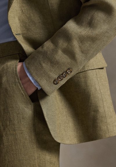 Mann trägt einen olivgrünen Anzug mit Struktur, vier braunen Knöpfen am Ärmel und eine Hand in der Tasche.