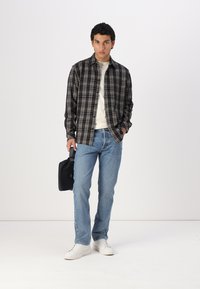 Black and white plaid shirt over a cream shirt, blue jeans, and white sneakers. Holding a black bag, standing on a white background.
