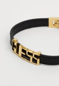 Svart lærarmbånd med et gullfarget metalllogo i fet skrift, festet med en gullklips. Glatt tekstur og minimalistisk design.