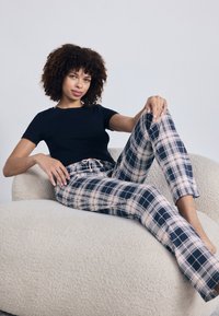 Haut noir à manches courtes ; pantalon à carreaux rose et marine avec taille à cordon. Texture douce, design ajusté et coupe décontractée. Assis sur un canapé de couleur claire.