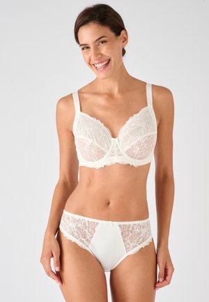 Donna sorridente che indossa un reggiseno di pizzo bianco e biancheria intima di pizzo abbinata su uno sfondo grigio uniforme.