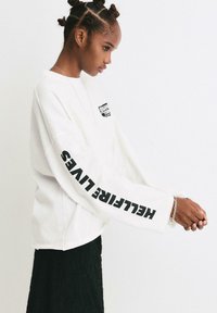 Bílé oversized mikina s dlouhými rukávy, s černým logem na hrudi a nápisem "HELLOFIRE LIVES" tištěným na levém rukávu.