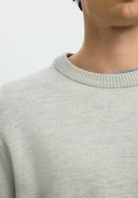 Artimas kaklo ir peties planas, vilkintis šviesiai pilką griovelio megztinį ant mėlyno marškinėlio prieš vientisą foną.
