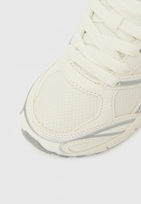 Pantof sport alb, cu partea superioară din plasă, panouri texturate și accente argintii. Dispune de șireturi plate și o talpă amortizată, axată pe confort.