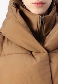 Giubbotto imbottito marrone con colletto alto, dotato di una chiusura a zip frontale e tessuto strutturato. Dettagli di cucitura visibili lungo le cuciture.
