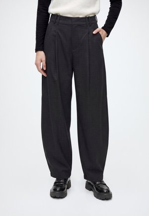 Personne portant un pantalon noir taille haute à jambes larges avec des plis, associé à des mocassins noirs épais, les mains partiellement dans les poches.