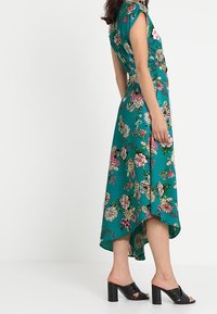 Femme portant une robe fleurie teal à volants asymétriques, avec des manches courtes et des sandales à talons noirs, se tenant devant un fond blanc.