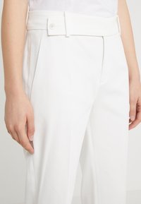 Zbliżenie na osobę noszącą dopasowane białe spodnie z zapięciem na guzik w pasie, ręce swobodnie opuszczone wzdłuż ciała.