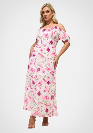 Lyshåret kvinde i hvid, ærmeløs maxikjole med pink og lilla blomsterprint, poserer med hænderne på hoften mod en ensfarvet baggrund.