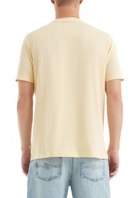 Man draagt een effen lichtgele T-shirt met korte mouwen en lichtblauwe denimjeans, gezien van achteren tegen een witte achtergrond.
