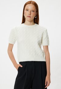 Suéter blanco de punto trenzado de manga corta con escote redondo y textura suave, combinado con pantalones negros.