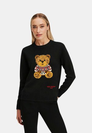 Kvinde iført en sort sweater med en teddybjørn-grafik i en rød, hvid og sort mønstret sweater, står med hånden i lommen.
