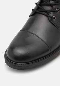Zapato de cuero negro con una textura suave, punta redondeada y detalles cosidos a lo largo de la suela. Presenta cordones negros y ojetes de metal.
