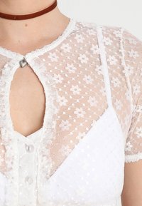 Weißes Spitzenoberteil mit floralen Mustern, einer vorderen Öffnung mit herzförmigem Akzent und kurzen Ärmeln mit gewelltem Saum.