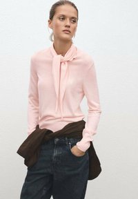 Maglione rosa chiaro a maniche lunghe con dettaglio del colletto annodato, abbinato a jeans scuri a vita alta e un indumento marrone legato in vita.