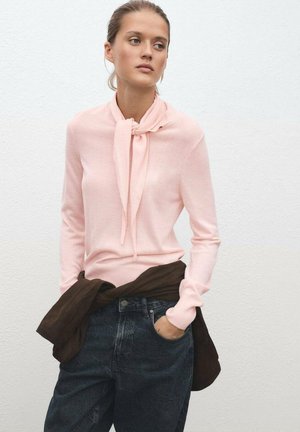 Pull en rose clair à manches longues avec un détail de col noué, associé à un jean foncé taille haute et un vêtement brun noué autour de la taille.