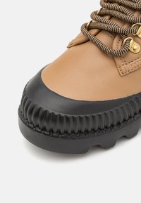 Botte en cuir tan avec semelle en caoutchouc noire. Présente des accents en caoutchouc texturé et des œillets dorés. Les lacets sont sombres avec un motif en zigzag.