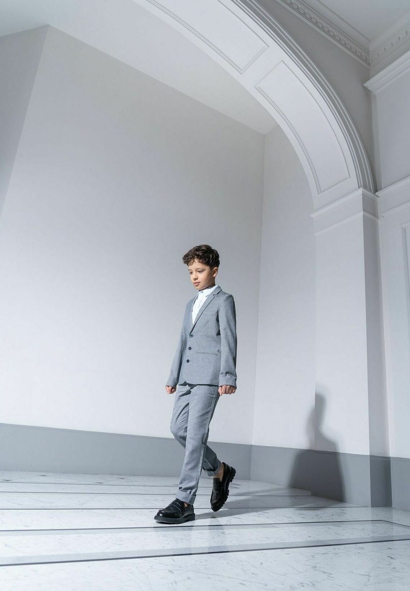 Szary garnitur z klapami w kształcie notcha, czarnymi guzikami i dopasowanymi spodniami. W parze z białą koszulą i czarnymi butami. Gładka tekstura, dopasowany krój.