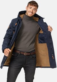 Doudoune marine avec une doublure beige chaude, fermeture éclair à l'avant et accents en épingle. Le modèle porte un col roulé noir et une ceinture marron avec un jean.