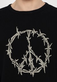Black ribbed long-sleeve shirt featuring a prominent white barbed wire peace symbol in the center. Soft texture, round neckline.