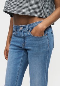 Sieviete tērpusies gaiši zilās auduma džinsu biksēs un melni baltā rūtaina īsa topa, ar vienu roku džinsu priekšējā kabatā.