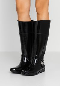 Czarne, lakierowane, wysokie botki do kolan o gładkim wykończeniu, z paskiem z metalowymi elementami i okrągłym detalem z logo po bokach.
