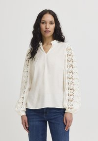 Blusa bianca con scollo a V e maniche a palloncino arricciate, caratterizzata da dettagli di pizzo intricati. Tessuto morbido, abbinata a jeans blu scuro.
