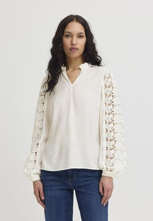 Vit blus med V-ringning och samlade ballongärmar med intrikata spetsdetaljer. Mjuk textur, i kombination med mörkblå jeans.