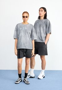 Sivi oversized majice s grafikom sigurnosne igle, crne traperice, i različite tenisice. Na jednom modelu sunčane naočale; verski akcenti.