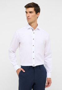 Camicia bianca a maniche lunghe con una vestibilità slim, caratterizzata da bottoni marroni e un colletto a fantasia. Abbinata a pantaloni blu navy.
