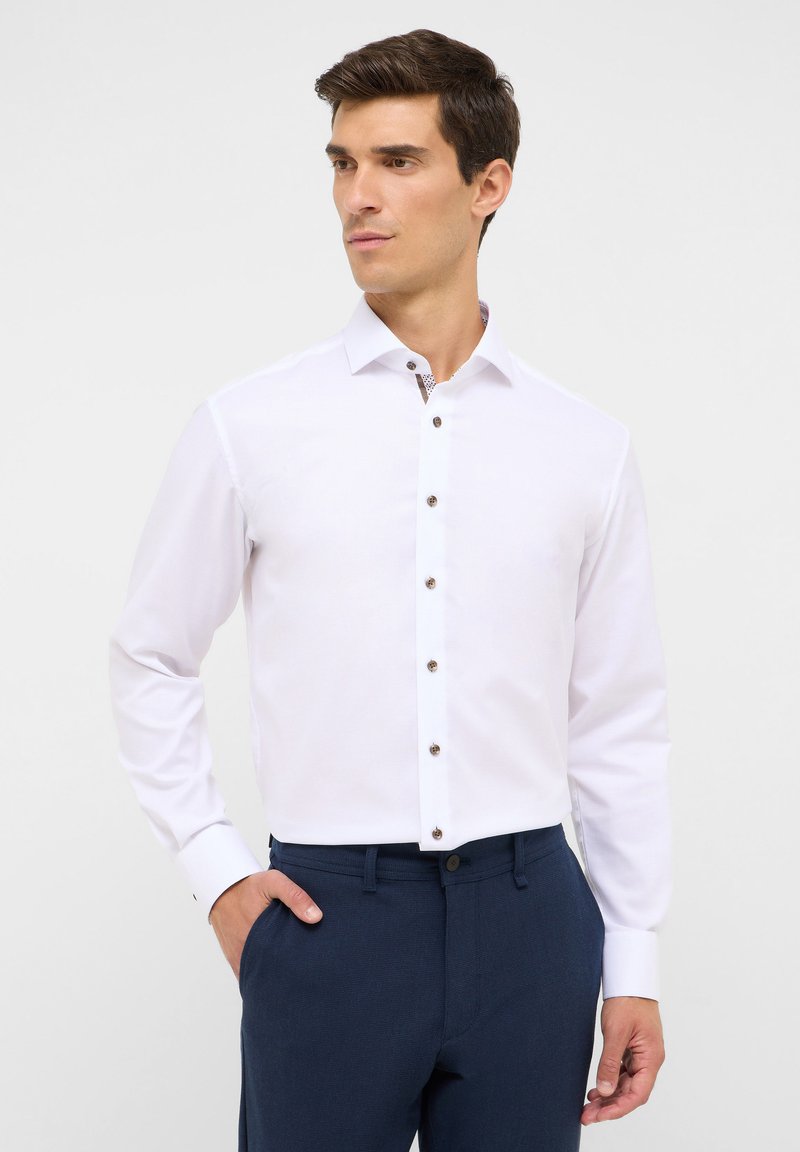 Camicia bianca a maniche lunghe con una vestibilità slim, caratterizzata da bottoni marroni e un colletto a fantasia. Abbinata a pantaloni blu navy.