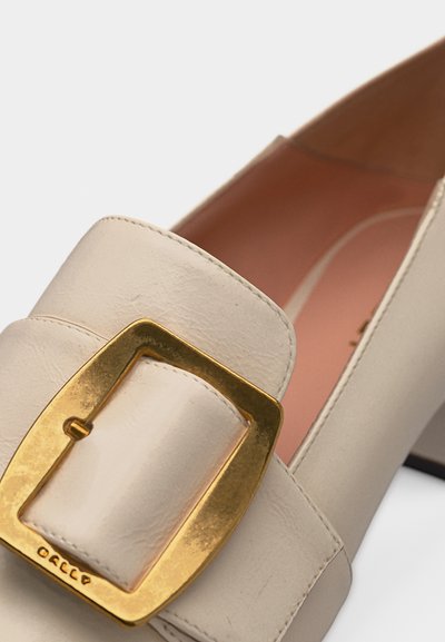 Chaussure à talon haut en cuir beige avec un grand détail de boucle carrée dorée et un bout arrondi, vue en gros plan.