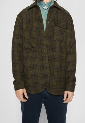 Mann trägt ein olivgrün-schwarz kariertes Hemd mit Knöpfen über einem helltürkisfarbenen Zip-up-Oberteil, kombiniert mit dunkelblauen Jeans.